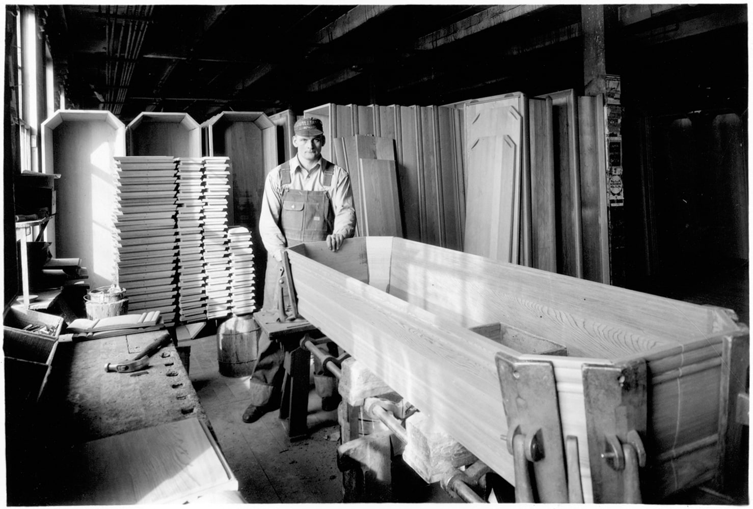 A Northwestern Casket Company worker standing at a bench beside a finished casket shell, with stacked lids and lumber behind him